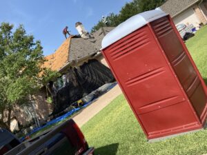 A Rust Red portable restroom trimmed out in white sitting on a lush green yard with a roof replacement in progress, provided by college station roofing services, Sustainable Roofing, for their contractors