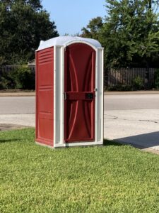 A Rust Red portable restroom trimmed out in white sitting on a lush green yard next to an empty drive way, provided by Sustainable Roofing for their contractors